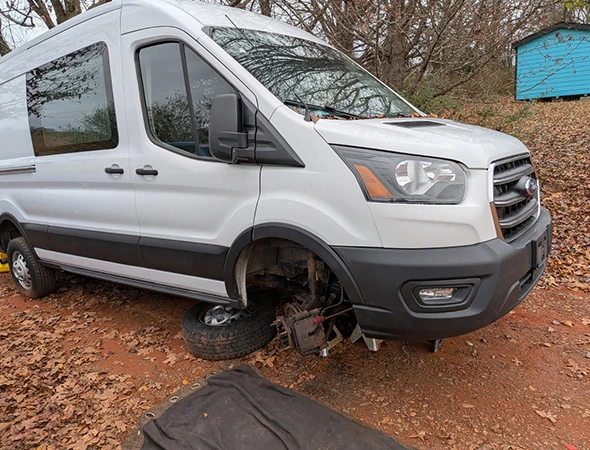 A white Ford Transit service van parked on a dirt/gravel surface with a front wheel removed for repair.