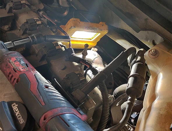 Mechanic working on a car engine cooling system under a work light, highlighting water pump repair services in Harrisburg, NC.